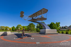 A stainless steel sculpture of the Wright Brothers and the 1903 Wright Flyer on display in the Huffman Historic District of Dayton under clear blue skies.