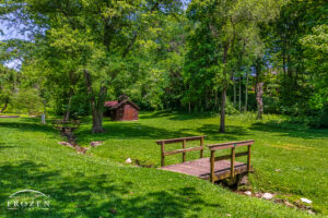 Bellbrook Park in Bellbrook Ohio on a pretty summer day where a foot bridge spans a creek that cuts through the park and leads the eye towards the park's sugar maple house