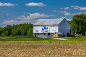 A white barn adorned with the Ohio Bicentennial Logo depicting a scene where the Coshocton barn basks in the summer sun will cumulus clouds floating in the sky