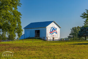 An ancient but sturdy white barn in the morning light as the sun illuminates the patriotic red, white and blue logo painted on its eastern side contrast nicely with the surrounding and lush green pasture