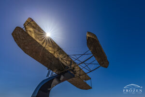 A stainless steel rendition of the Wright Flyer III mounted high on a pedestal where the background entails clear blue sky and a crisp sunburst.