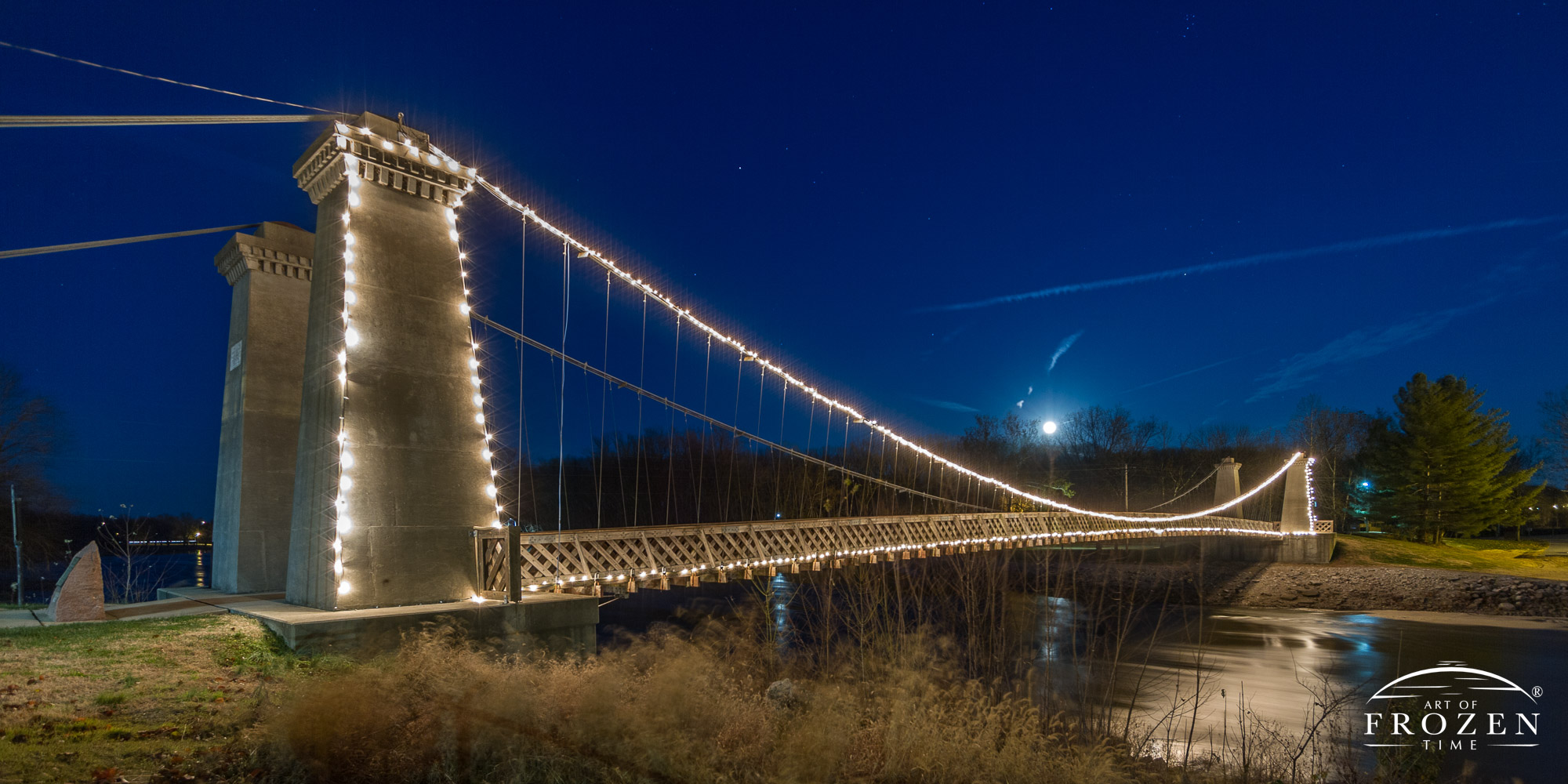 General Dean Bridge outlined in Christmas Lights as the Kaskaskia River flows underneath and moon rises in the blue night sky.