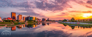 Panoramic Dayton Ohio skyline at sunset reflected in the glassy Great Miami River beside RiverScape MetroPark and the five-sided riverside pavilion.