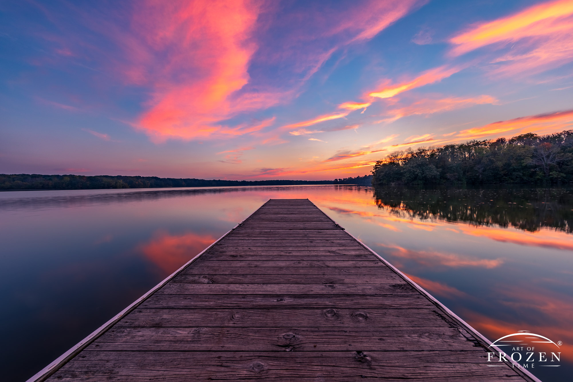 A boat dock leading the eye to the horizon where cirrus clouds bask in orange colors of twilight and calm lake waters reflect the sky above
