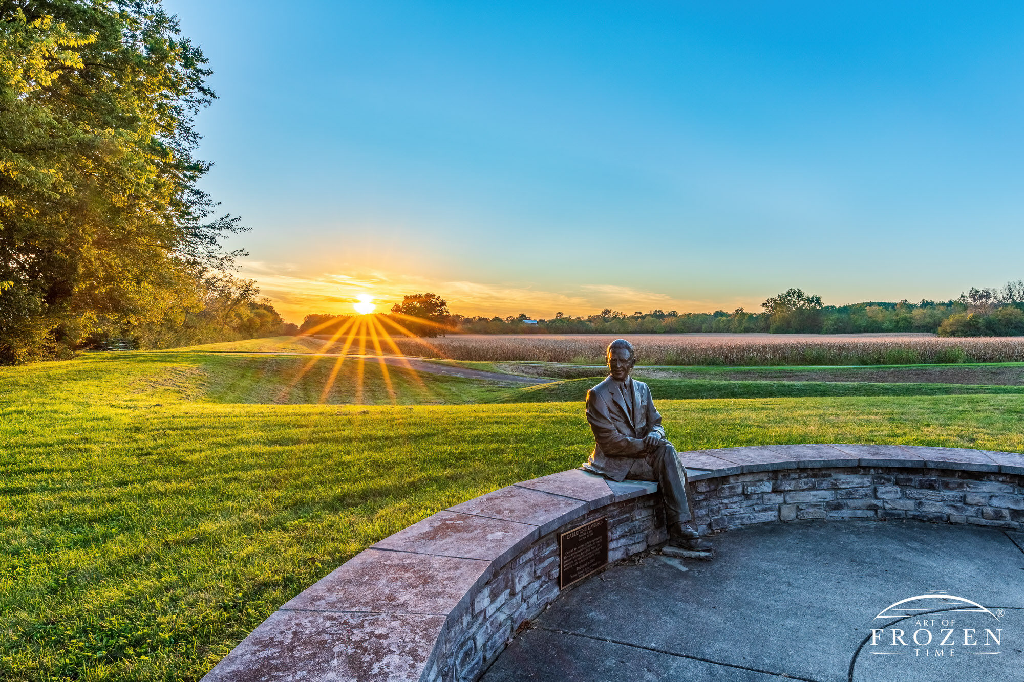 A bronze sculpture of Carleton F. Davidson sitting on a small wall during a fall sunset that cast golden light over the historical education center holding his name