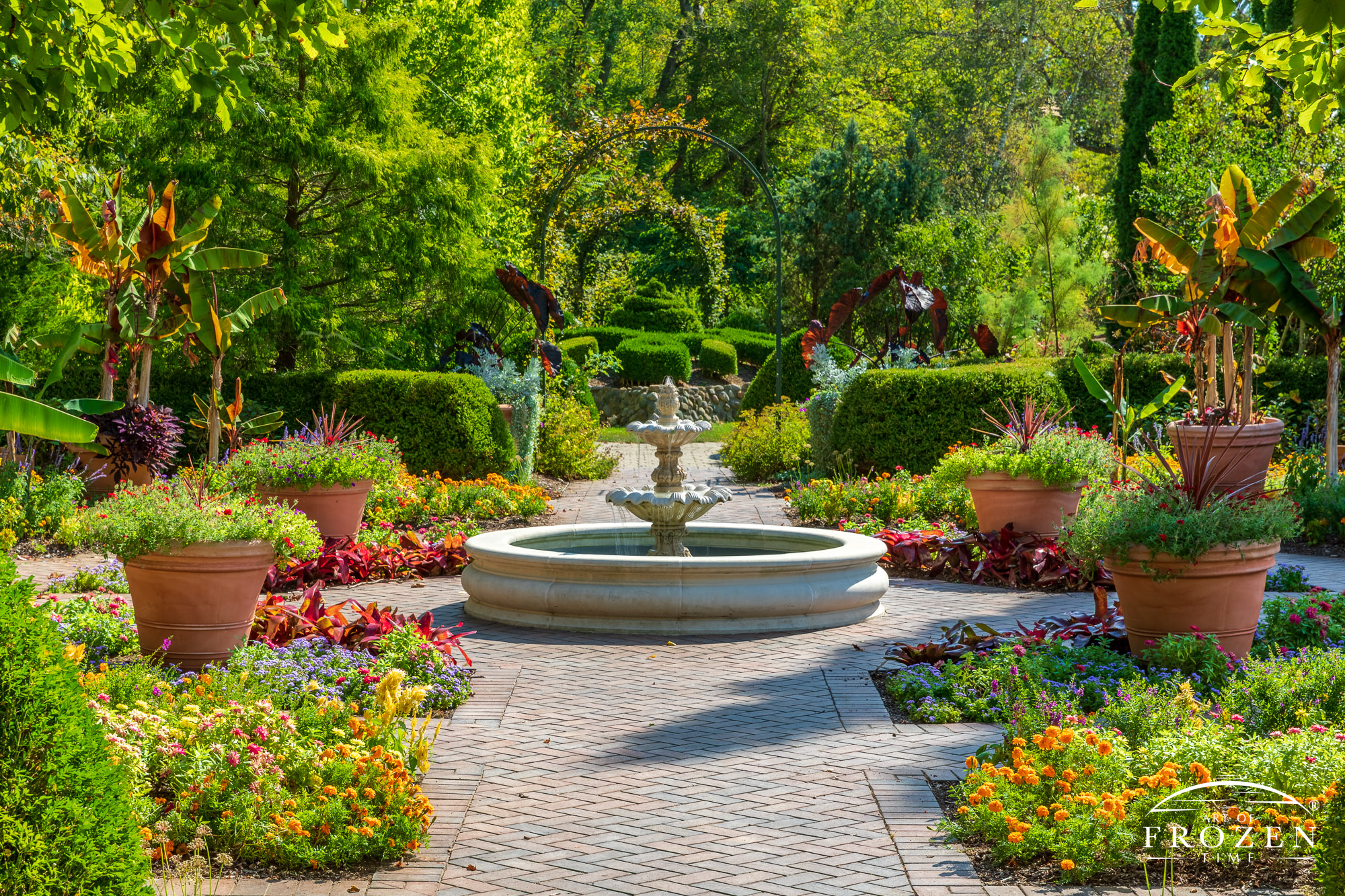 A formal English garden featuring a trickling fountain surrounded by lush and vibrant flowerbeds, pots and topiaries in the evening light.
