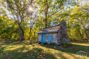An original Butler County log home resides in a wooded park on this autumn evening as the thinning leaves filter warm light from the setting sun.
