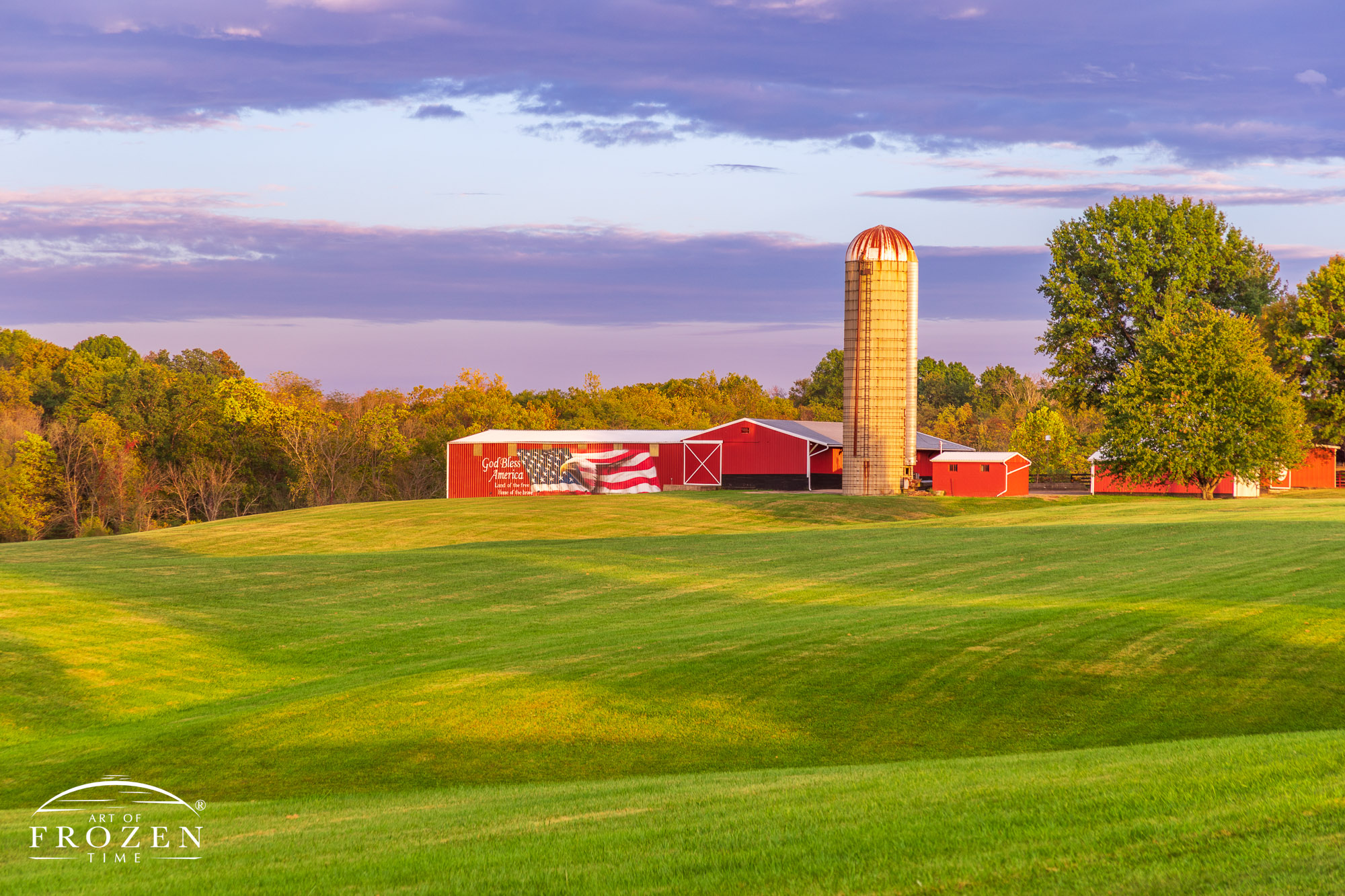 An evening view of an Ohio farm surrounded by rolling grass covered hills where every building is red with white time and one pole barn expresses the family’s patriotism