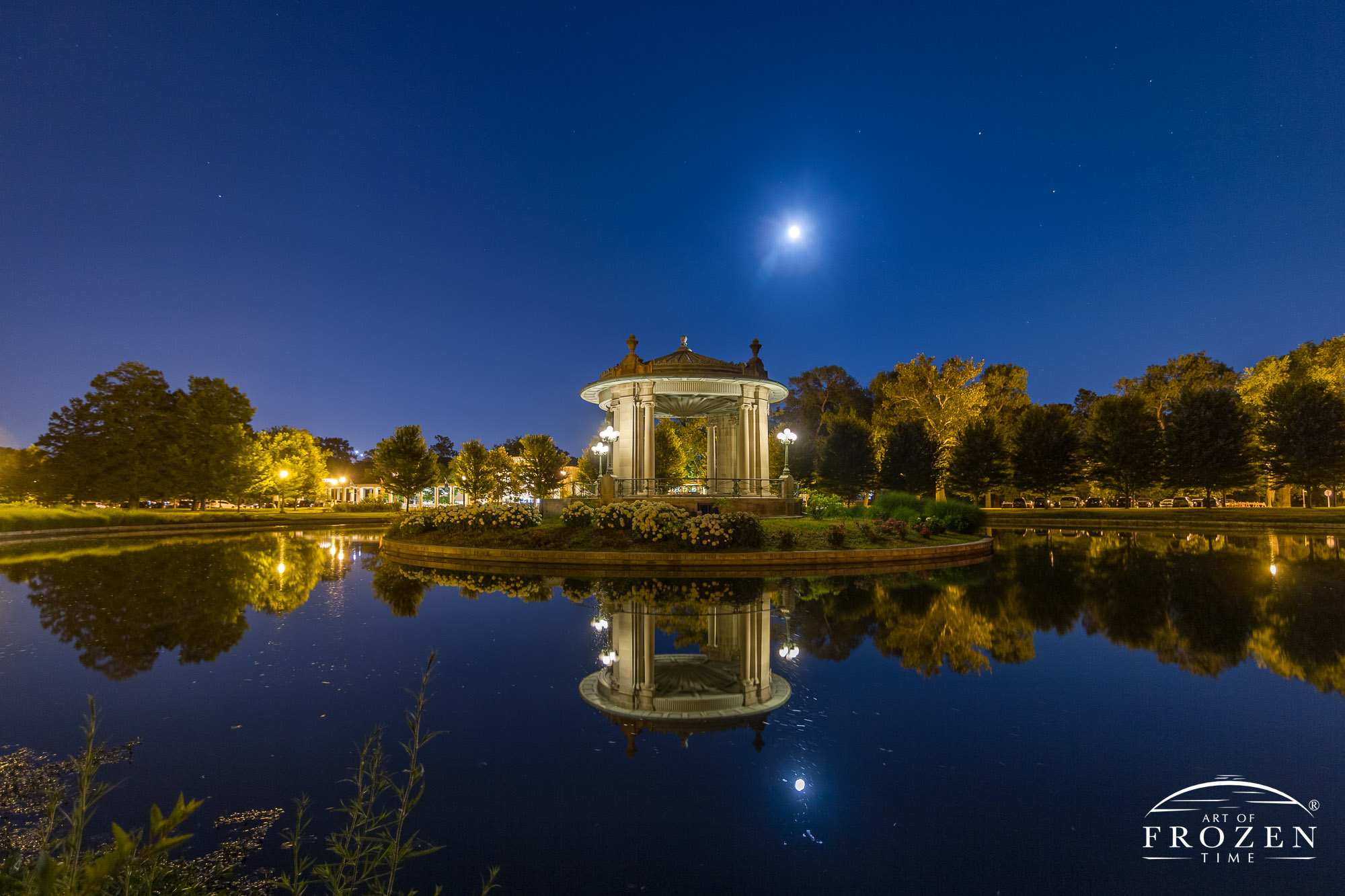 A nightscape of the Forest Park Nathan Frank Bandstand, in St Louis Missouri where a full moon hangs over the park and Pagoda lake reflects the sky above