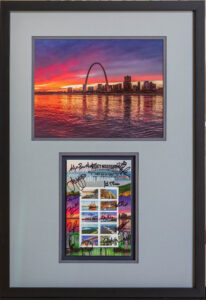 A framed print of a first day of issue stamp with signatures of attendees from the first-day ceremony matted with the original print of the St Louis Skyline