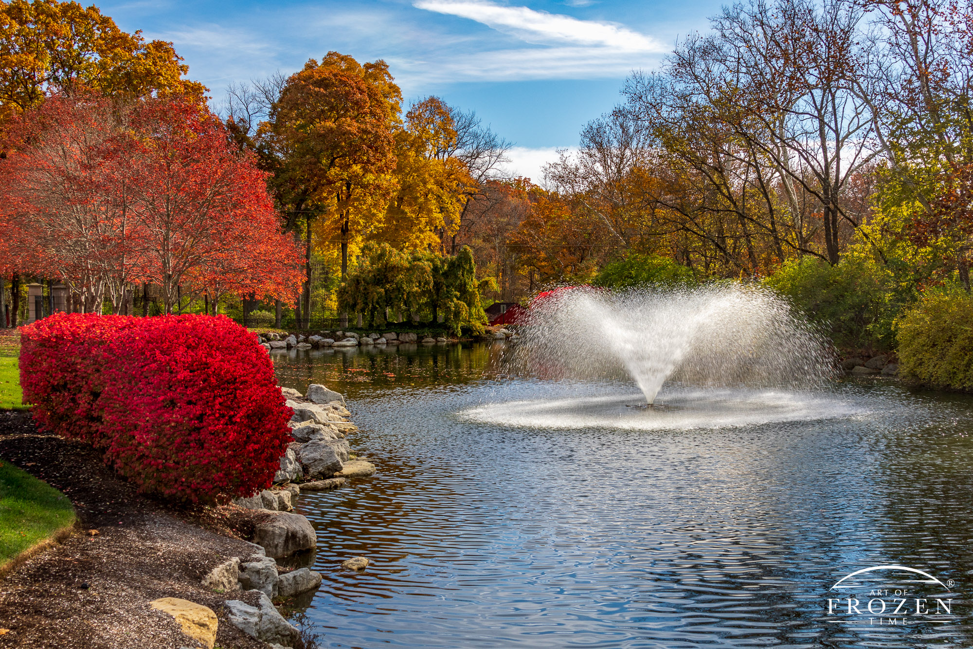 A fountain's spray glistens in the sunlight on a fall day where the surrounding vegetation glows in reds and yellows.