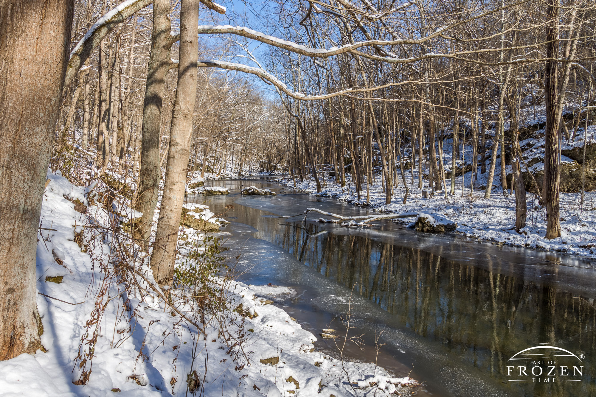 The Little Miami River gently flows by ice and thick snow as it passes through Clifton Gorge under blue skies and bright sunlight