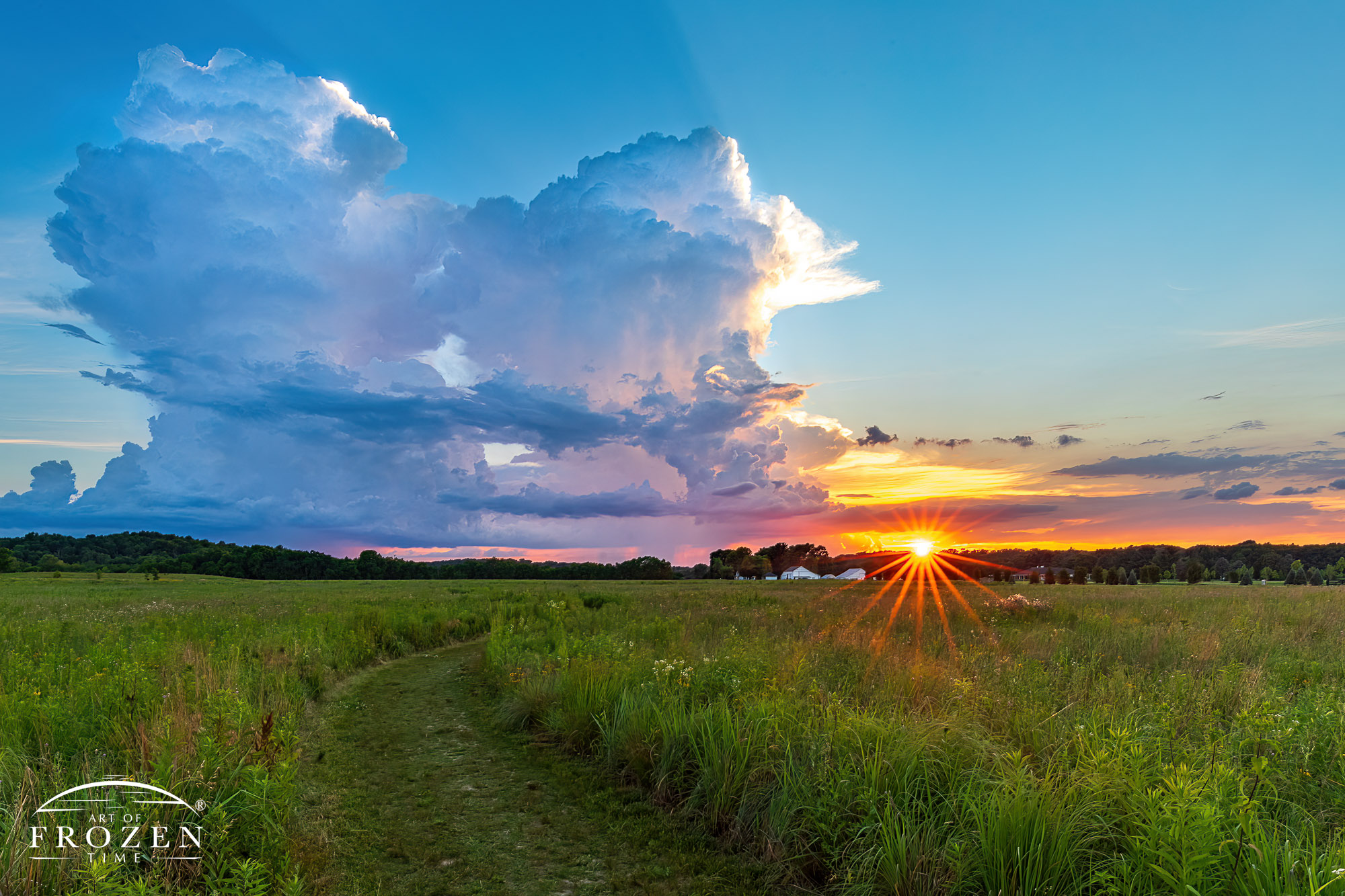 A prairie trail leads the viewer’s eye to the sunburst on the horizon and two cumulonimbus clouds which fill the western sky