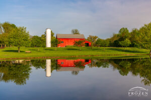 A traditional Ohio barn with metal roof, red paint and white silo next to a pond whose still waters form a perfect reflection