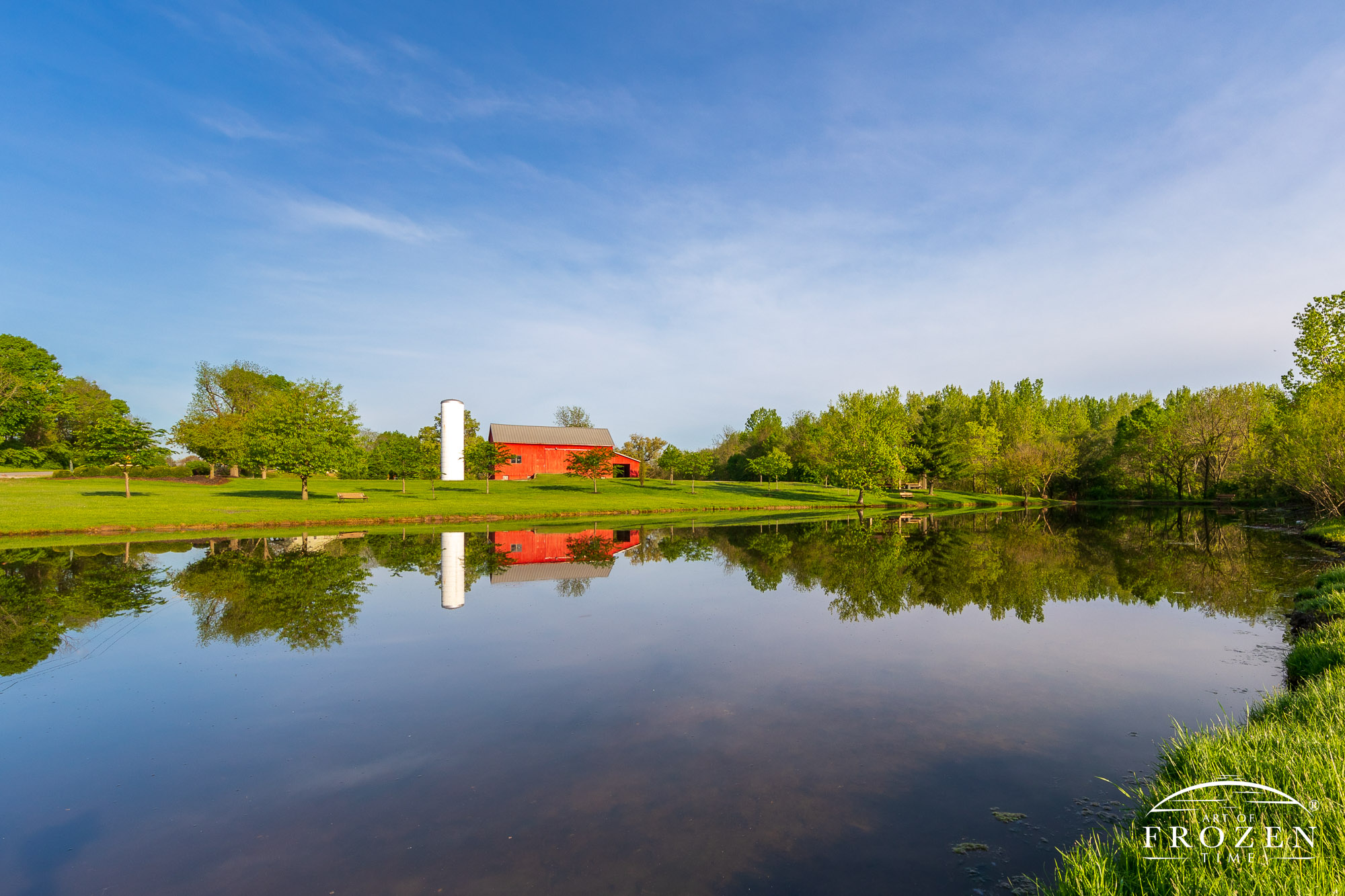 A traditional Ohio barn with metal roof, red paint and white silo next to a pond whose still waters form a perfect reflection
