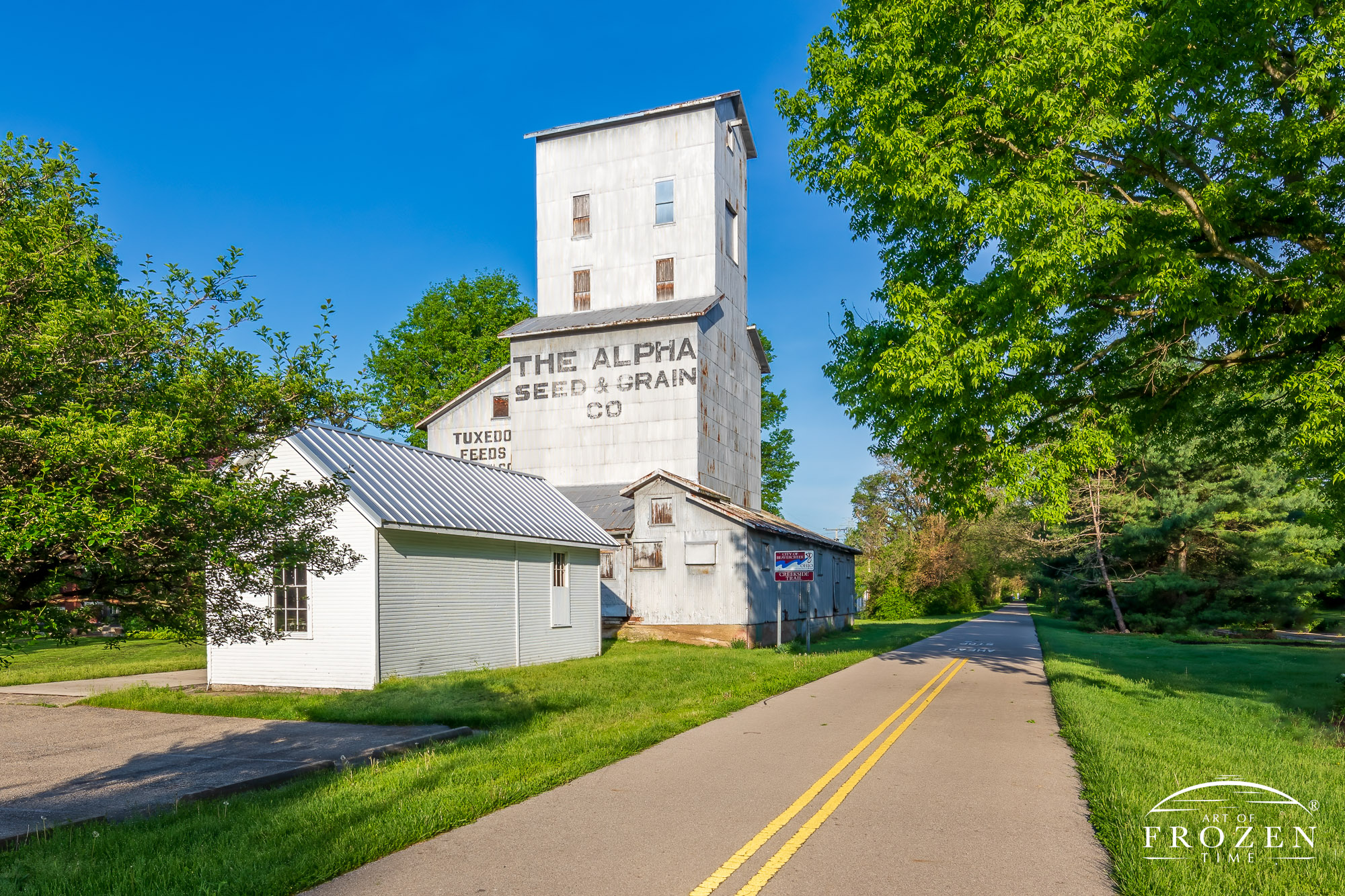An old grain elevator resides next to a bike path which runs off to the distant horizon as lush green vegetation lines the path lying under crystal-clear skies