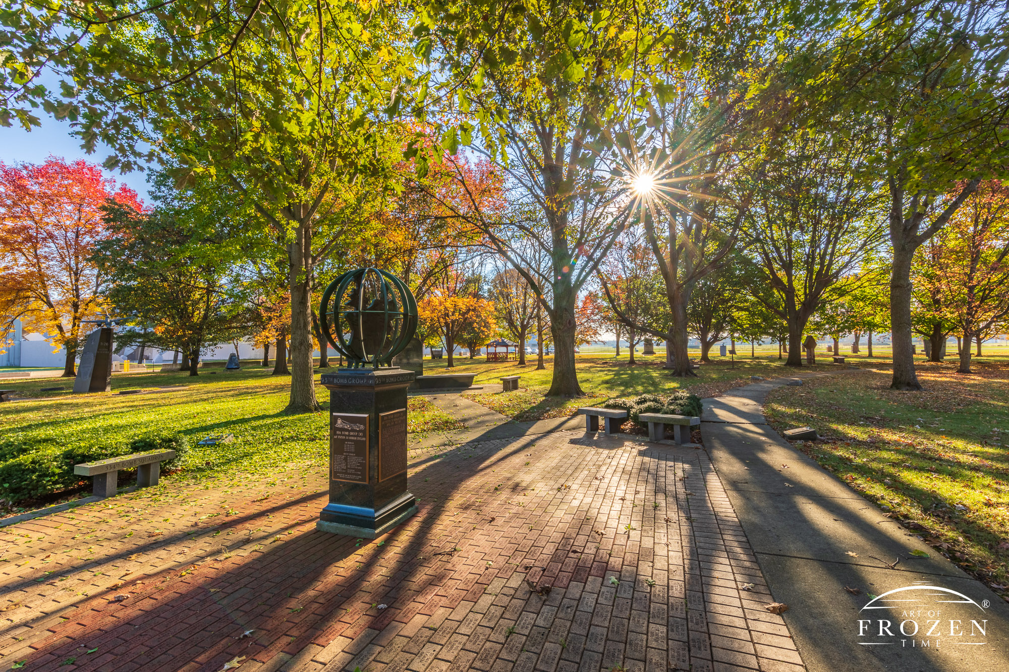 A memorial in the shape of a bronze globe standing along a brick paver sidewalk on an autumn morning where the early light rakes across the ground and backlights the colorful trees.