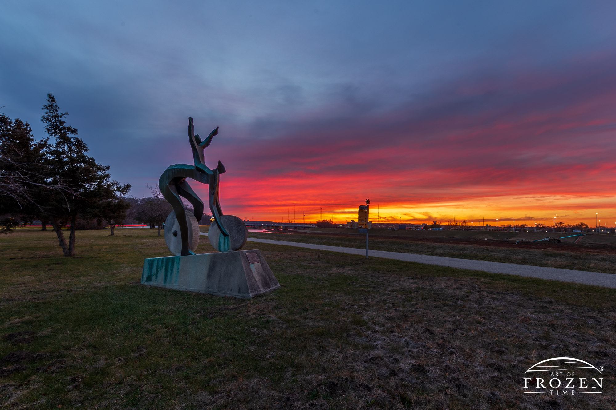 A modern sculpture celebrating bicycling stands along the Great Miami River Trail during a richly colored sunset. The rider’s outstretched arms strike joyous pose as if expressing excitement over the brief and brilliant display of color along the Dayton Ohio skyline.