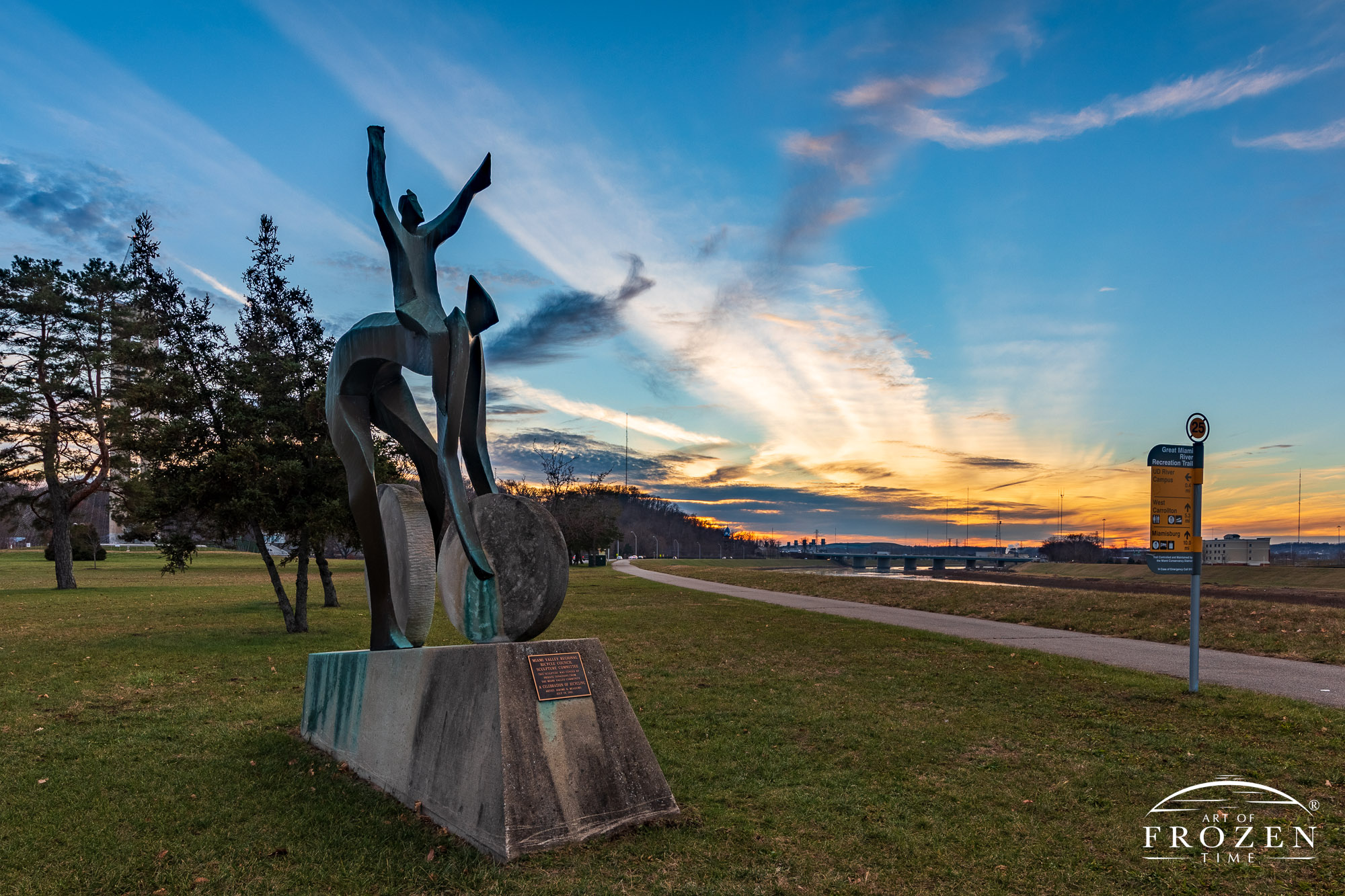 A sculpture which celebrates bicycling shows a human form riding handsfree with arms stretched skyway in this wispy sunset image along the Great Miami River trails