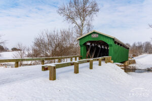 A green covered bridge stands about covered snow fields and under blue skies.