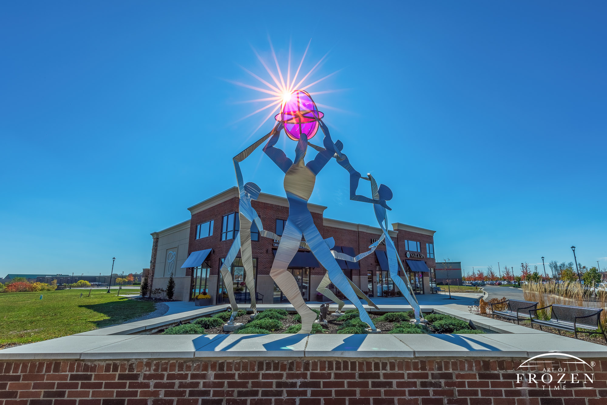 A stainless steel sculpture called Celebration lies in Cornerstone Park, Centerville Ohio as the sun backlits the stained glass ornament being held in the air by a stylized family dancing in a circle.