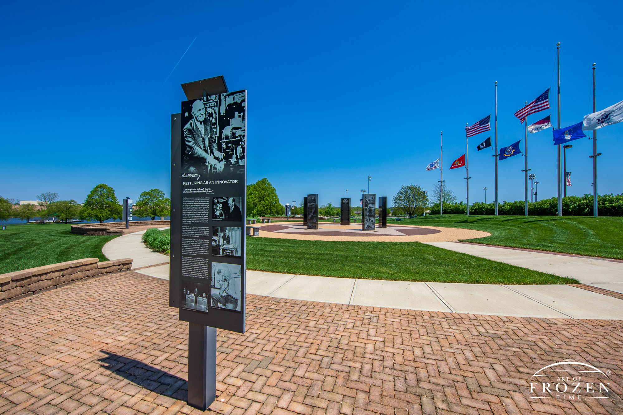 A scene from Delco Park that captures the Charles F. Kettering History Walk and the Kettering Veterans Memorial on a gorgeous spring day where the flags flap in the gentle wind under blue skies.