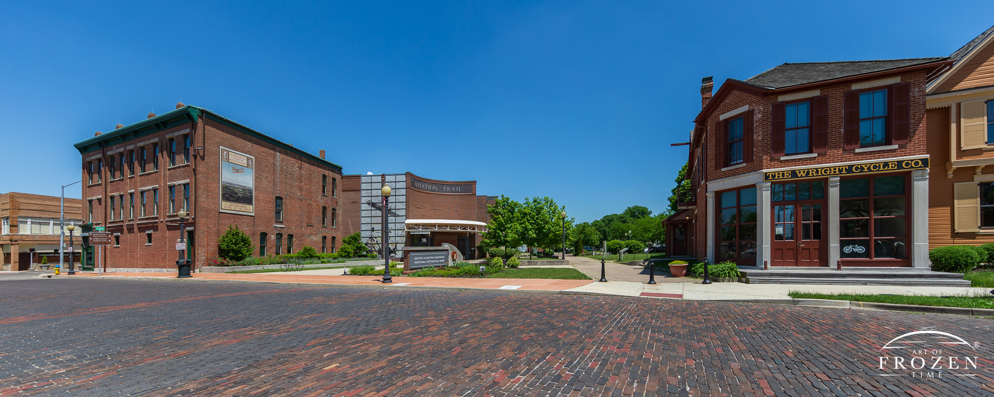 A panorama Image featuring a replica of the red-bricked Wright Cycle Company and Wright-Dunbar Interpretive Center under clear blue skies.