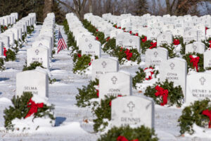 A patriotic image featuring orderly rows of white military headstones with each marker having its own green wreath and red ribbon, all covered in a fresh blank of snow on Christmas morning