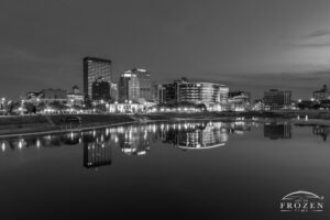 A black and white image of twilight over the Dayton Skyline where the glassy Miami River relects the city lights and evening sky