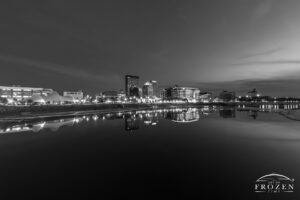A black and white image of twilight over the Dayton Skyline where the glassy Miami River relects the city lights and evening sky