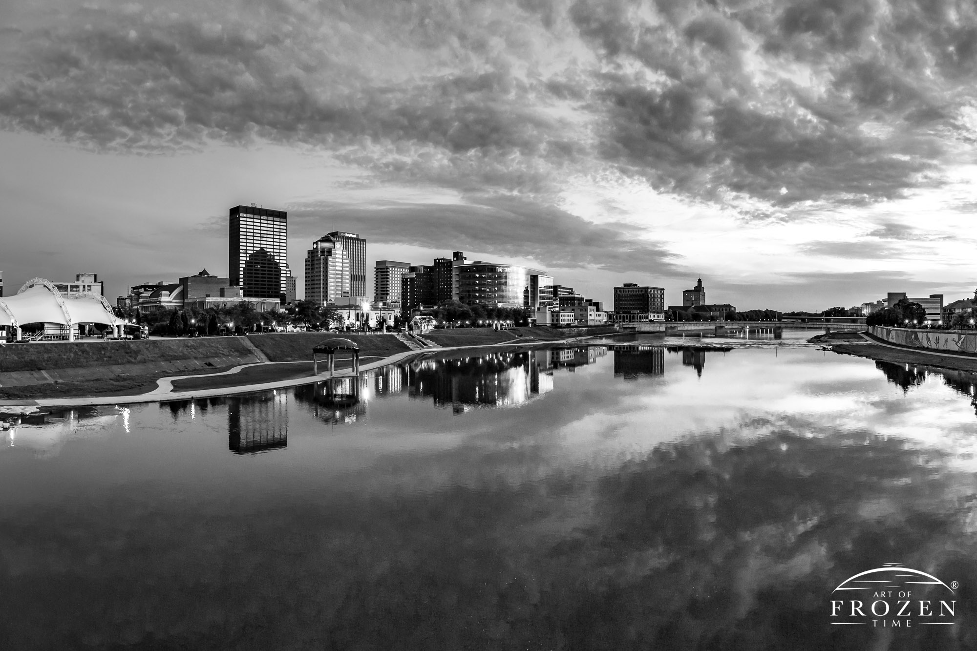A black and white image where the sun shines the Dayton Skyline and the office windows and smooth river reflect the contrasting light as intersting clouds fill the sky.