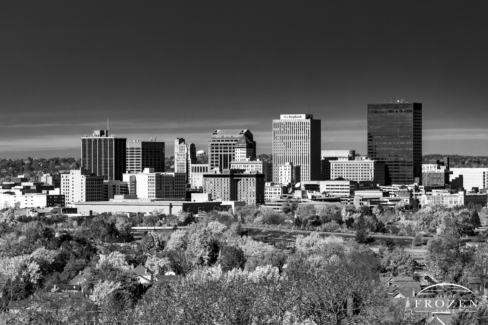 A black and white image of the Dayton Skyline from the east on a sunny n day where the colroful foreground trees reflect the autumn light as the blue sky takes on dark hues.
