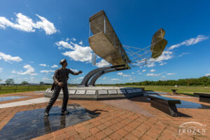 A stainless replica of the 1909 Wright Flyer resides on a pedestal outside Wright Patterson AFB as if it just launched into flight on a clear day with a few cumulus clouds