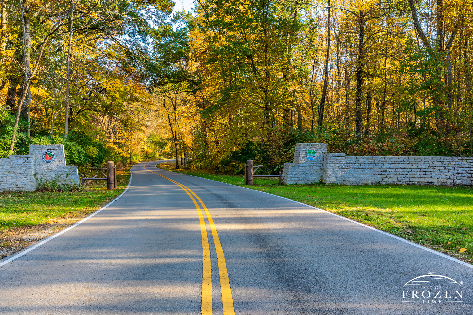 The road to John Bryan State Park extends into the distance under yellow sugar maple leaves as it passes by the stone entrance