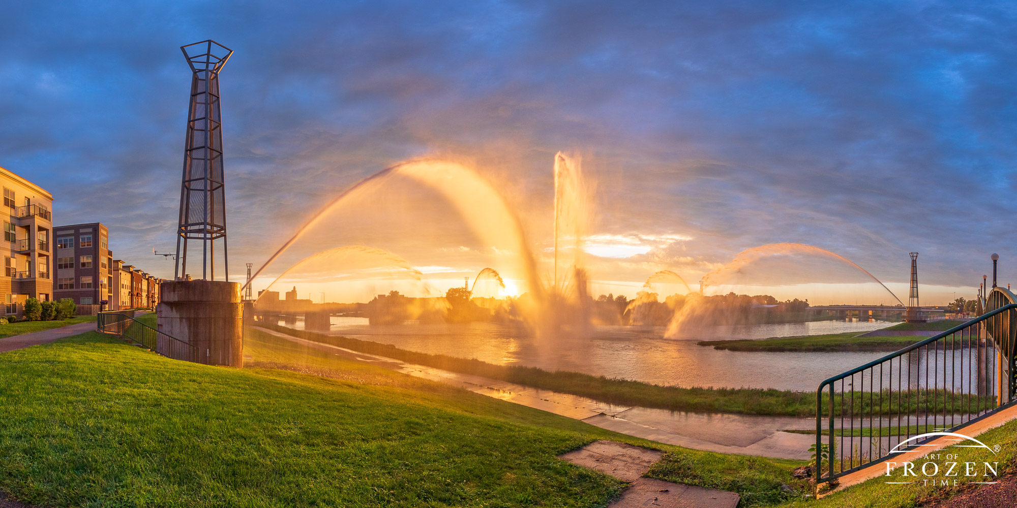 Dayton Ohio’s Fountain of Light erupts during a sunset where the sun peaks through the overcast sky and warmly illuminates the fountain spray on this summer evening