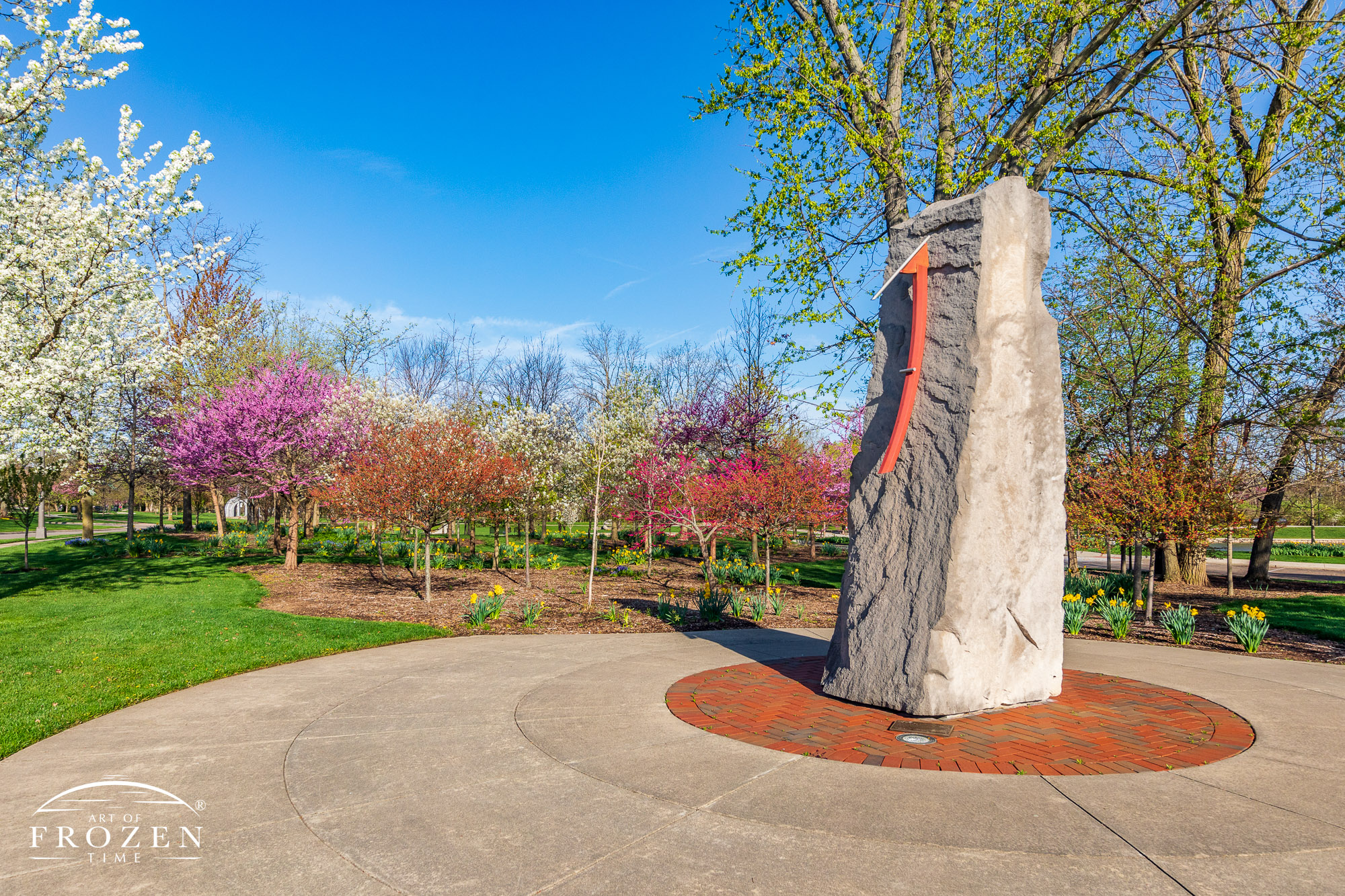 A vertical sundial mounted to a large stone which resides in a small plaza surrounded by blooming trees and daffodils.