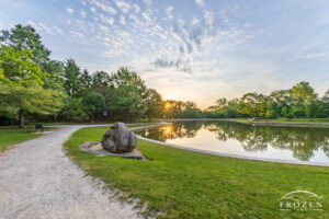 A sunrise at Kettering Ohio’s Lincoln Park where a pathway curves around a stone sculpture as the adjacent pond waters reflect the sun briefly creating two suns