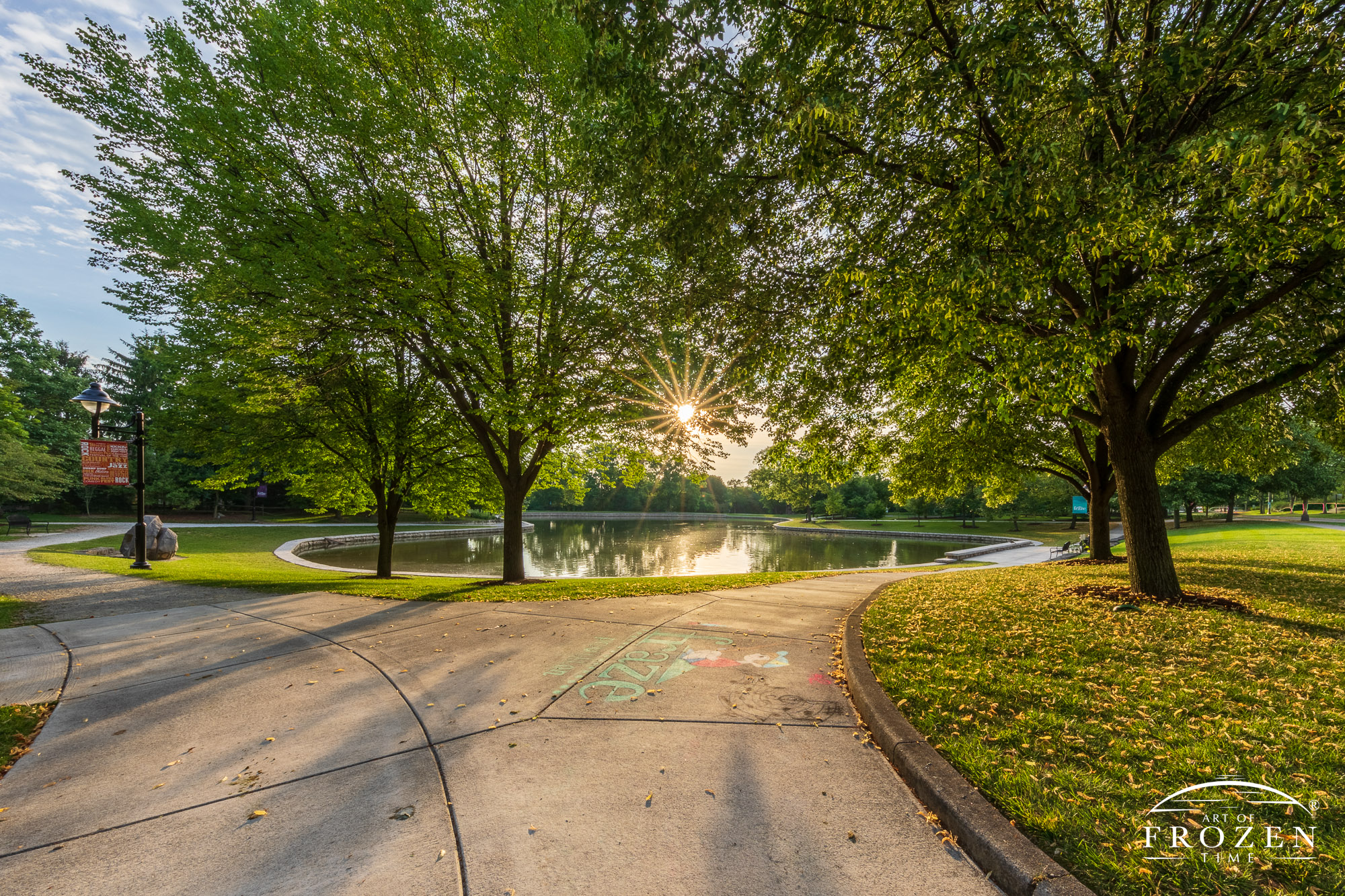 A pathway leads to a sunrise scene where the golden light illuminates Lincoln Park’s Pond and the surrounding trees filter the sunlight