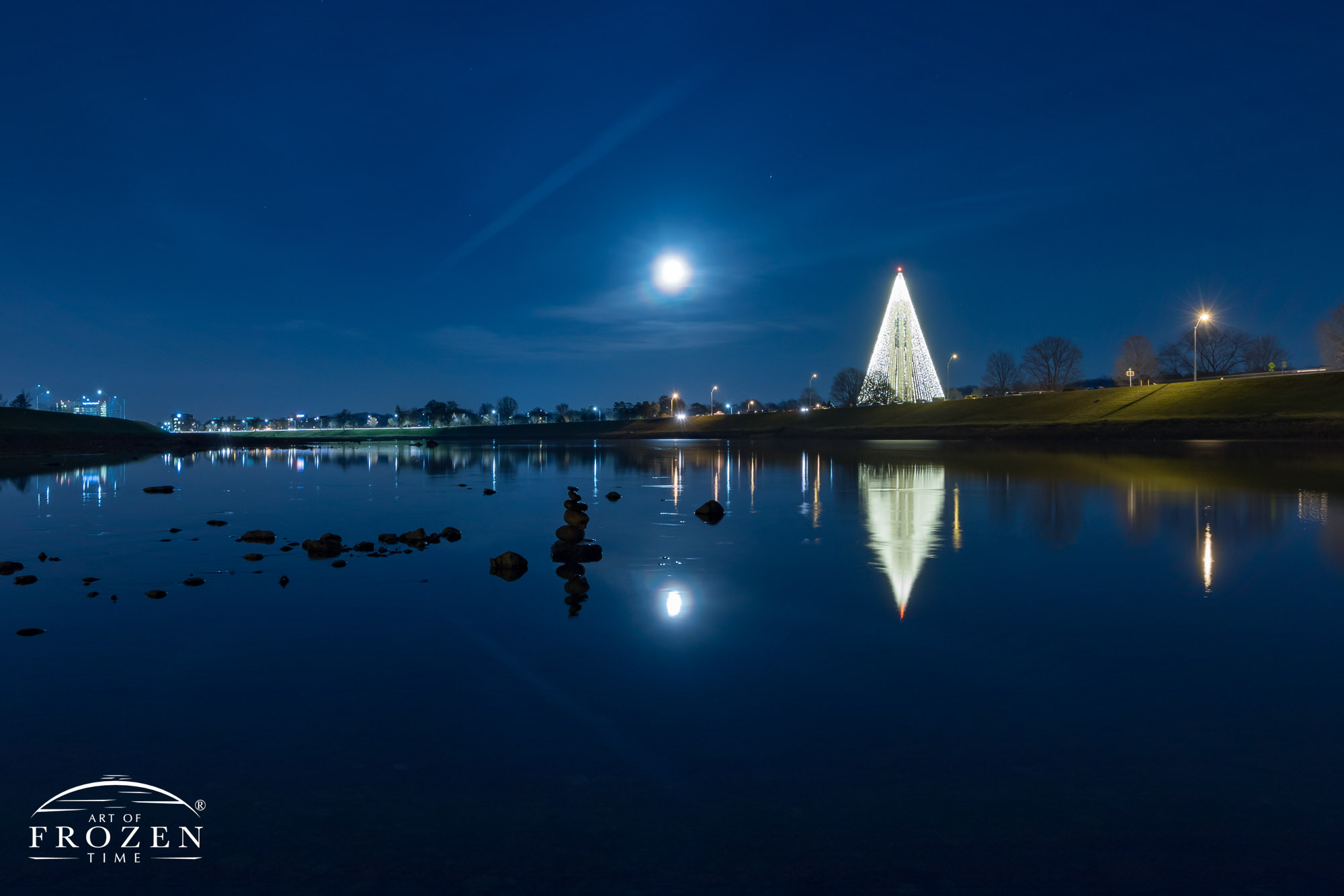 A nightscape where Deeds Carillon Tree of Light reflects in the still waters of the Great Miami River as the December full moon takes to the sky