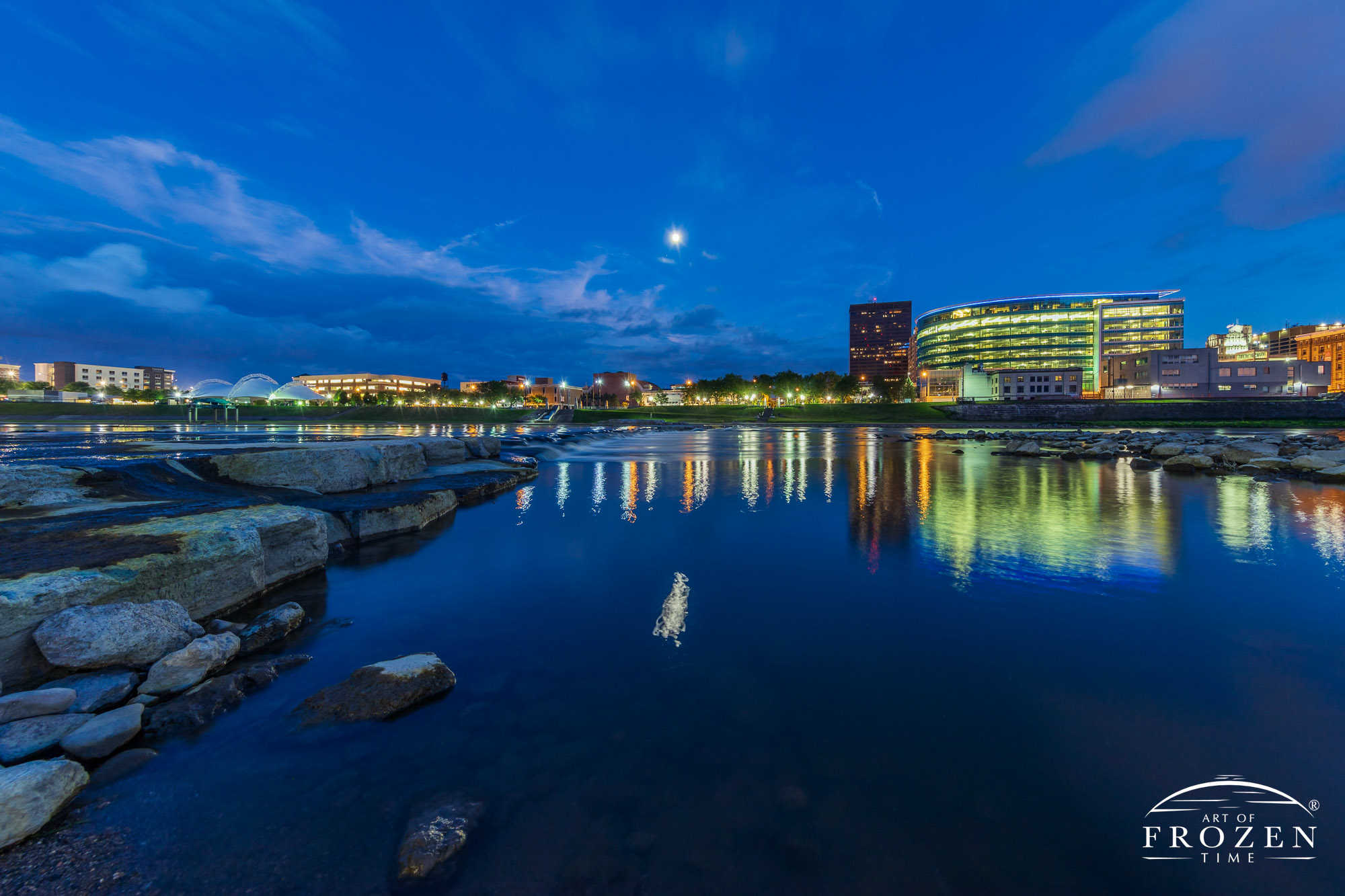 A twilight image of RiverScape’s RiverRun where the kayak chutes lead the eye to the opposite shoreline and the warmly lit CareSource Building all under the nighttime glow of a moon