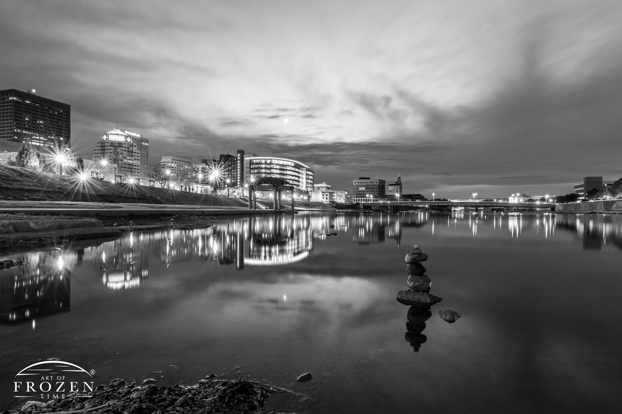 An evening scene of the Dayton Skyline rendered in black and white where the smooth Miami River surface reflects the RiverScape MetroPark lights.