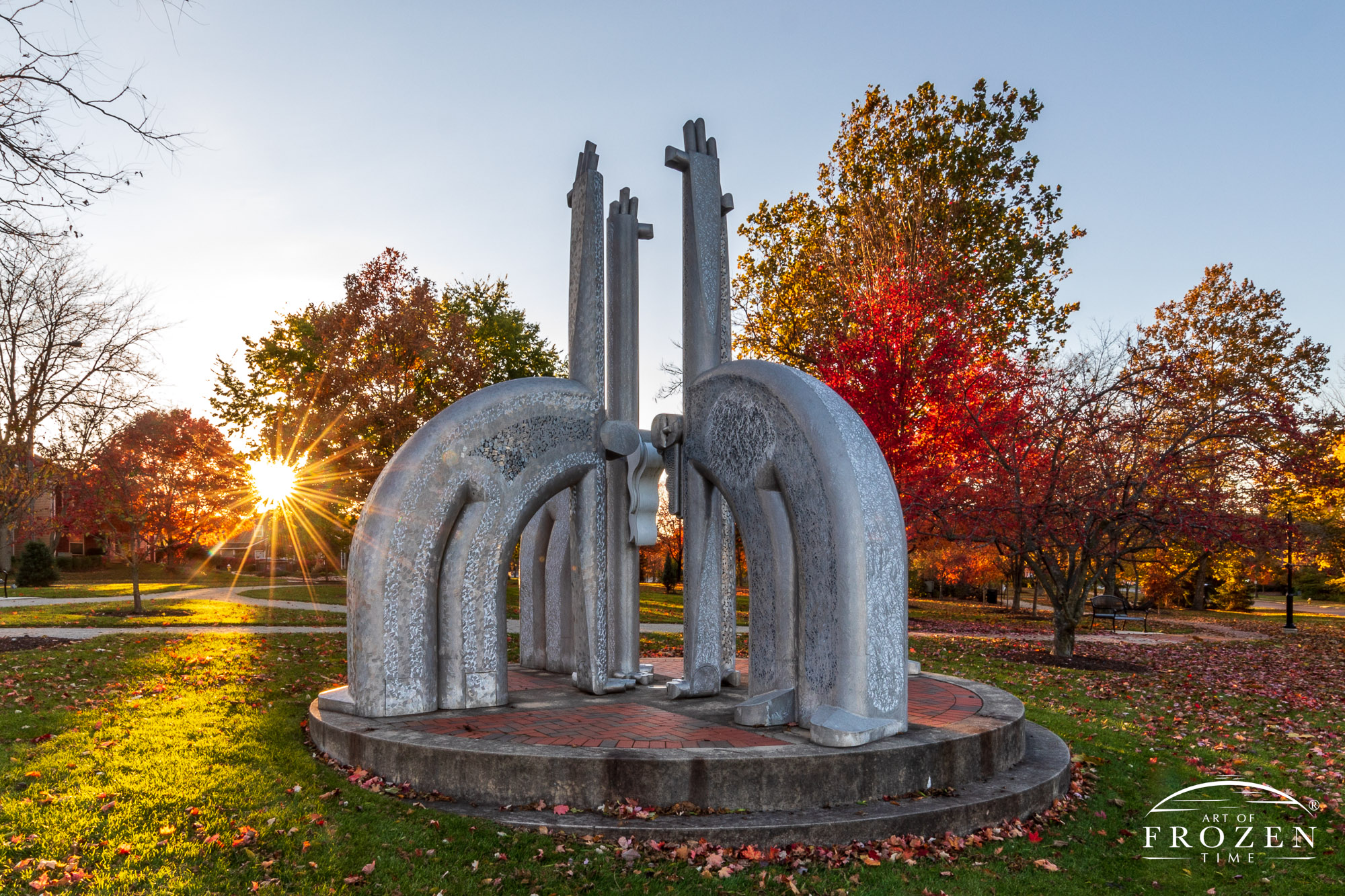 A fanciful aluminum sculpture with stylized figures reaching skyward, surrounded by autumn-colored trees backlit by the setting sun.