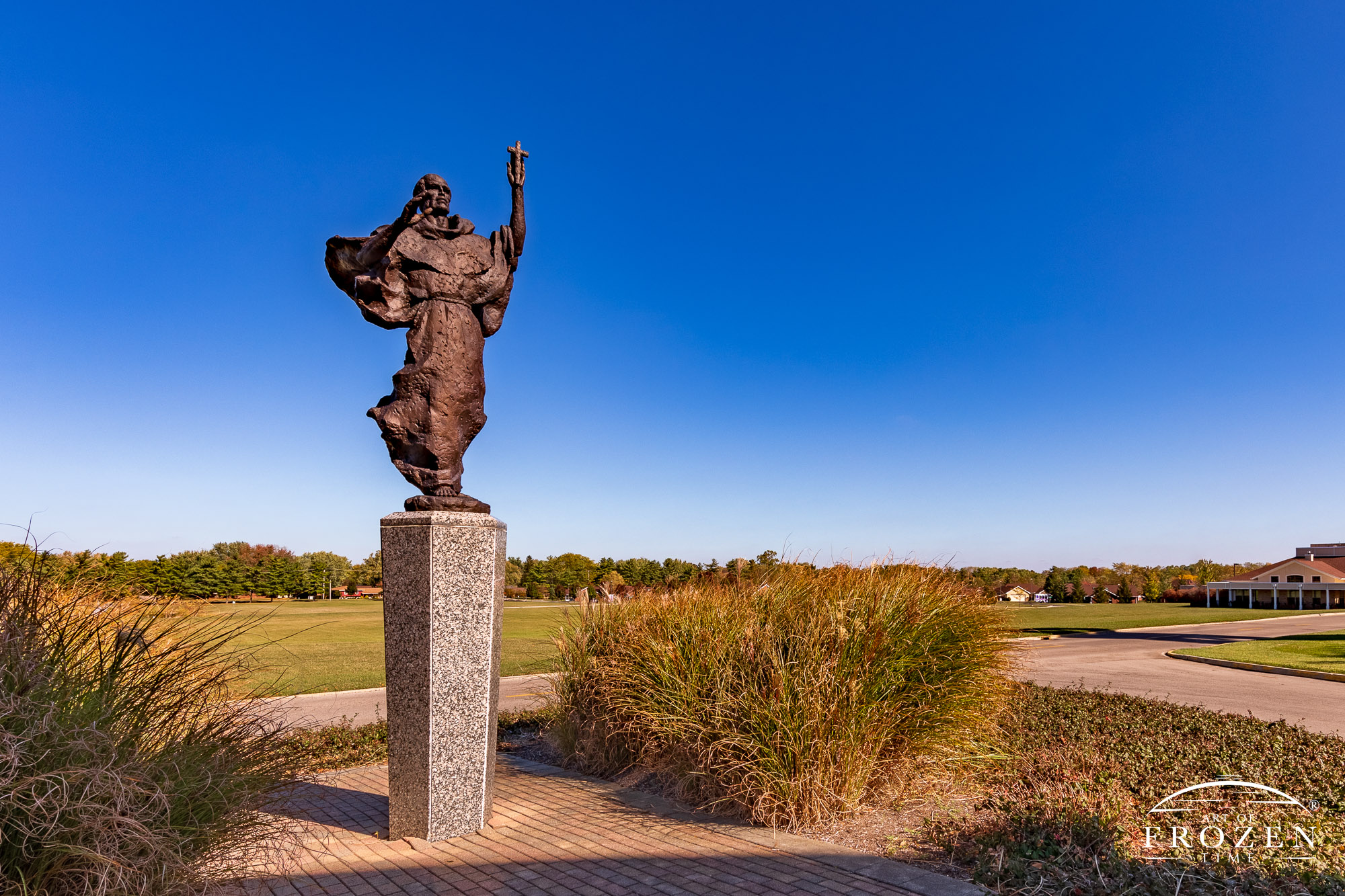 A bronze sculpture of St. Francis of Assisi holding a crucifix while staring skyward which reside under blue azure skies and golden light