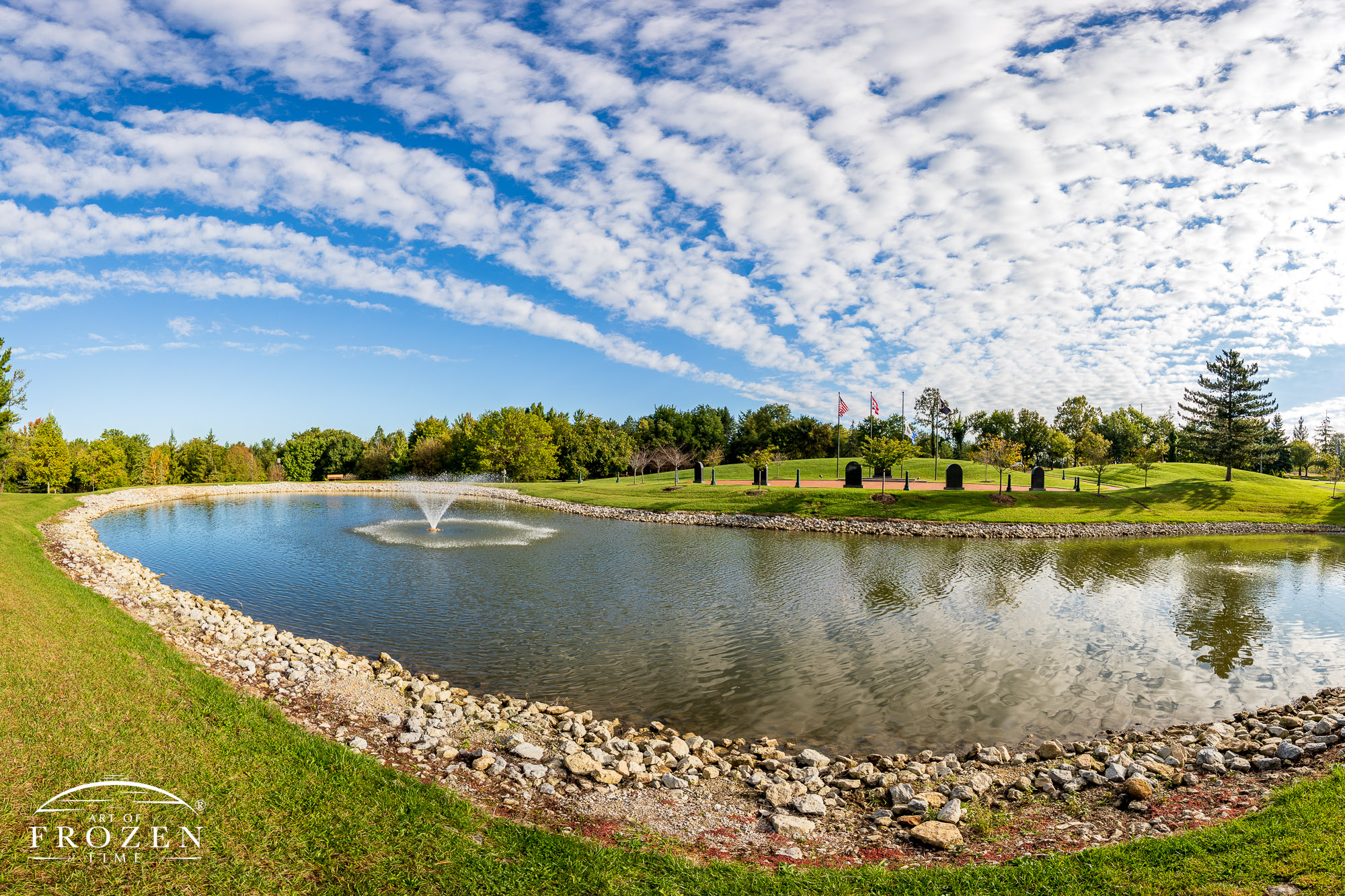 A wide-angle view of the fountain and pond at Stubb Park as band of clouds stretch across the blue sky