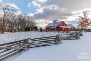 A snowy scene where a stacked split-rail fence zig zags to the red barn structure which serves as the Carriage Hill MetroPark visitors center.