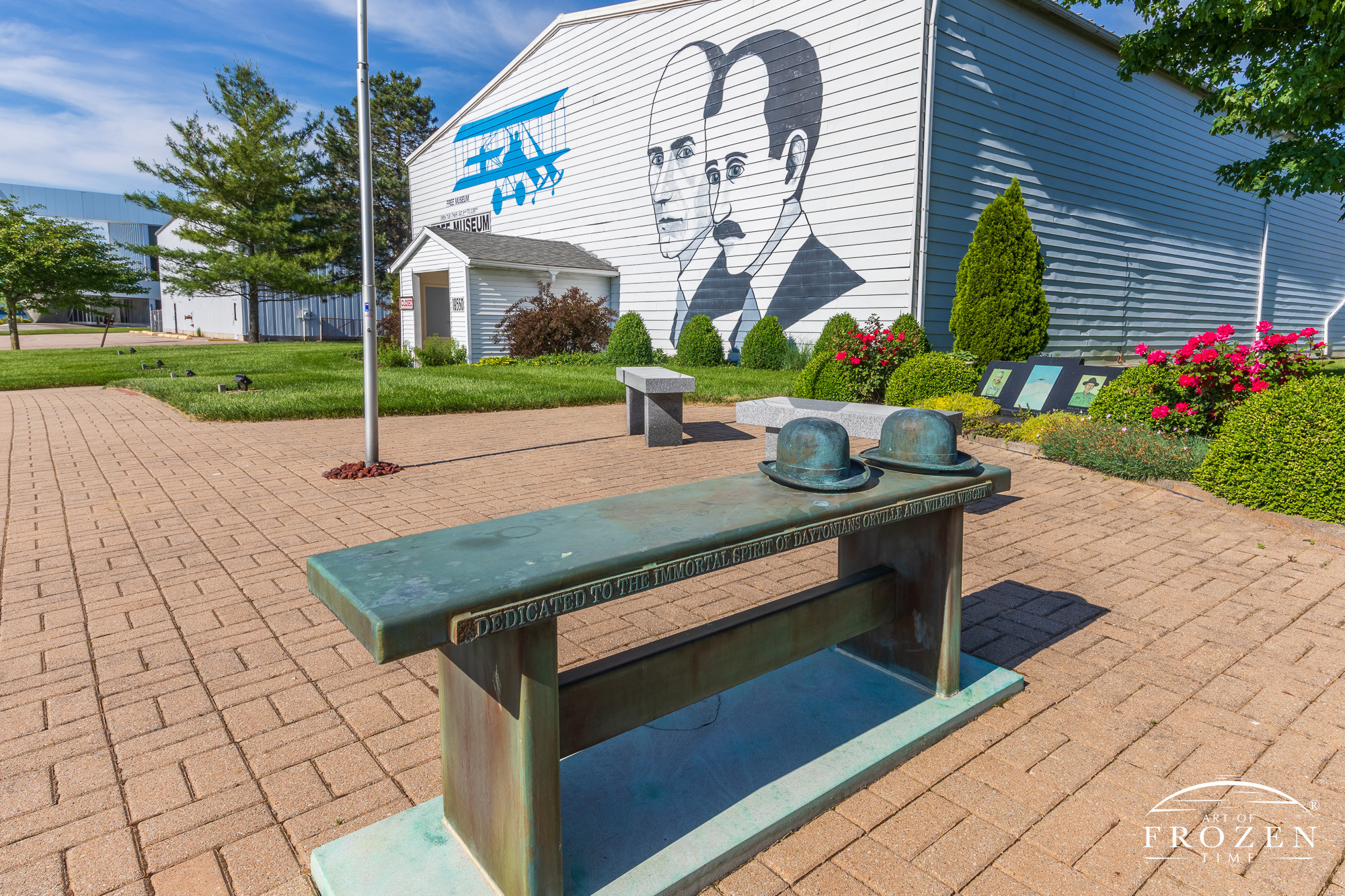 Mural of the Wright Brothers and their Wright B Flyer on a June evening while a bronze bench sculpture featuring their classic derby hats sits in the foreground