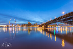 twilight view of the Eads Bridge as it crosses the Mississippi River towards St Louis where the calm river reflects the bridge, skyline and Gateway Arch.