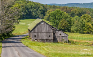 An unpainted barn with a green roof in Pennsylvania as a country road bends around the structure and over rolling hills of Brackney PA