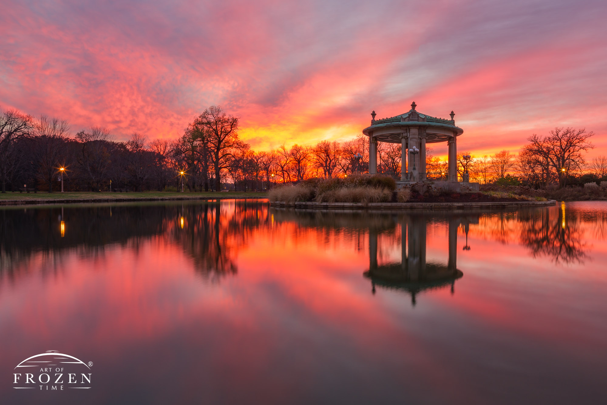 The St Louis Forest Park Nathan Frank Bandstand during a colorful sunset where Pagoda Lake reflects the sky and the silhouetted bandstand