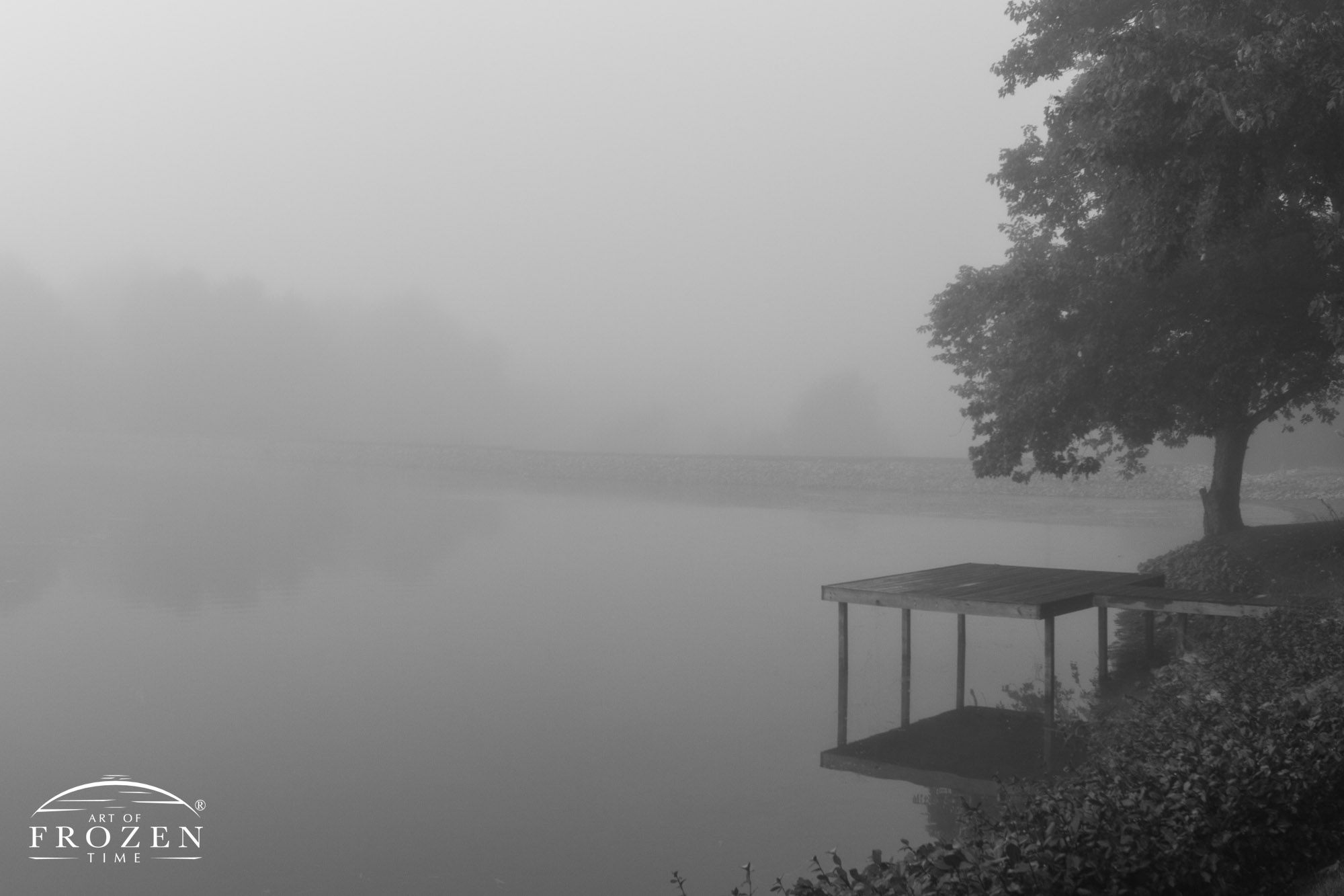 A dock leading to a large pond whose distant shoreline is obscured by heavy fog making for a moody image over O’Fallon Illinois.